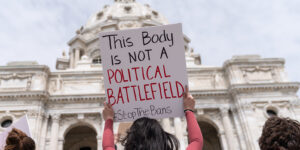 A woman standing in front of the US Capitol, surrounded by other women, holding a sign that reads: "This Body Is Not A Political Battlefield #StopTheBans"