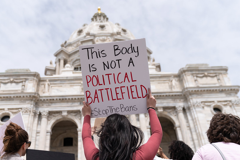 A woman standing in front of the US Capitol, surrounded by other women, holding a sign that reads: "This Body Is Not A Political Battlefield #StopTheBans"