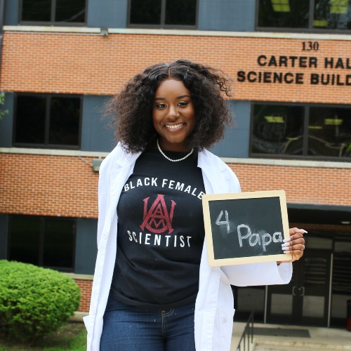 Hailey wearing a t-shirt that says "Black Female Scientist" standing in from of her campus science building, holding a sign that says "4 Papa."