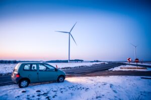 A car driving on a road on a snowy evening in front of a wind turbine.
