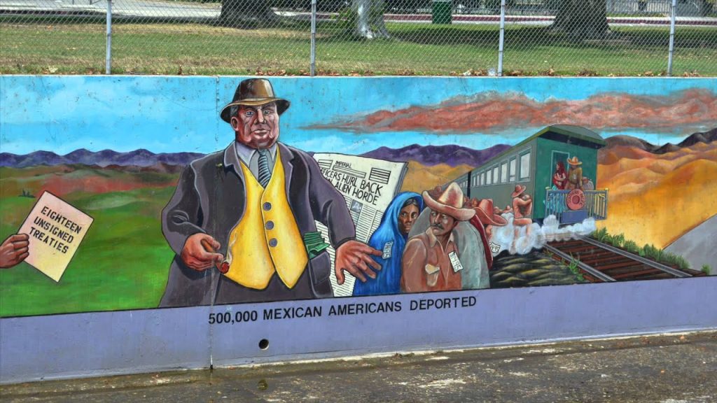 A mural painted underneath a chain link fence of a line of Mexican Americans reluctantly boarding aa train while a man in a fancy suit and pockets full of cash pushes them onwards. A hand on the left hand of the mural holds a document labeled "Eighteen Unsigned Treaties". Underneath the mural is written the words: "500,000 Mexican Americans Deported."