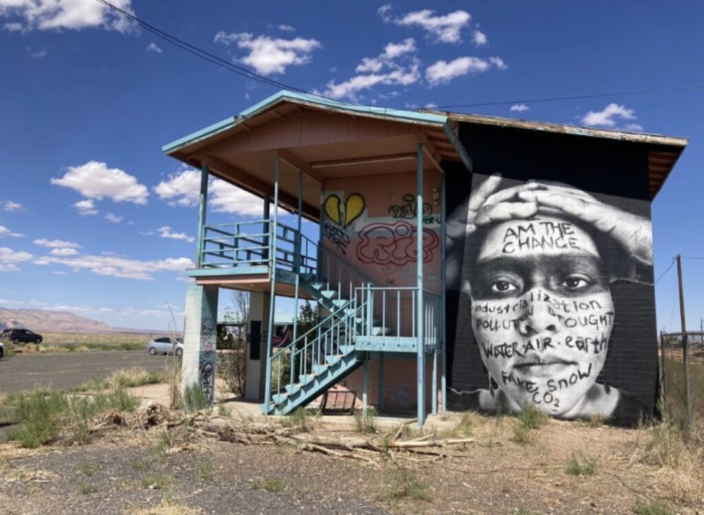 A photograph of a Native American person placed on the wall of a small building as a mural. On the person's face is written the words: "I am the change, industrialization, pollution, drought, water, air, earth, fake snow, co2.