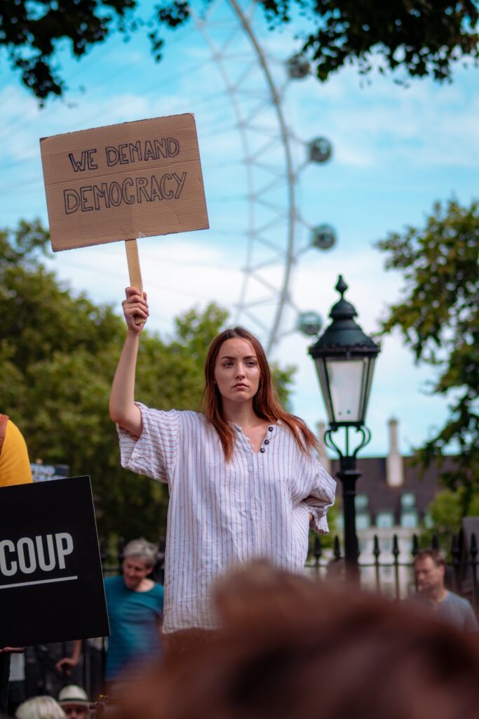 Woman standing in front of a crowd holding a sign that reads: "We Demand Democracy"